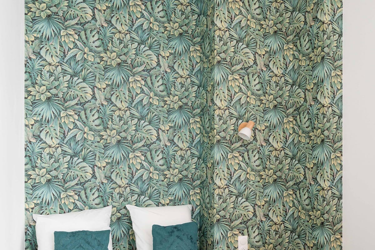 Una cama bien hecha con ropa de cama blanca y verde azulado, una mesita de noche de madera con una lámpara y un papel tapiz con estampado de hojas tropicales.