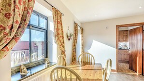 A dining area with a wooden table and chairs, a window with floral curtains, and a view of a balcony. - Betty Hutton - UK36661 (Belchford)