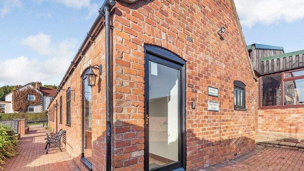 A brick building with a black door and a small window. There is a bench outside and a sign on the wall. - Betty Hutton - UK36661 (Belchford)
