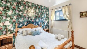 A bedroom with a floral wallpaper, a wooden bed frame, a bedside table with a lamp, and a window with curtains. - Betty Hutton - UK36661 (Belchford)