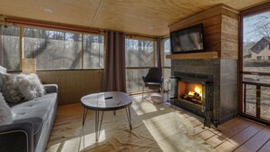 A cozy living room with a fireplace, a sofa, a TV mounted above the fireplace, and large windows. - Cozy Waterfront Escape near Helen & Vineyards (Cleveland)
