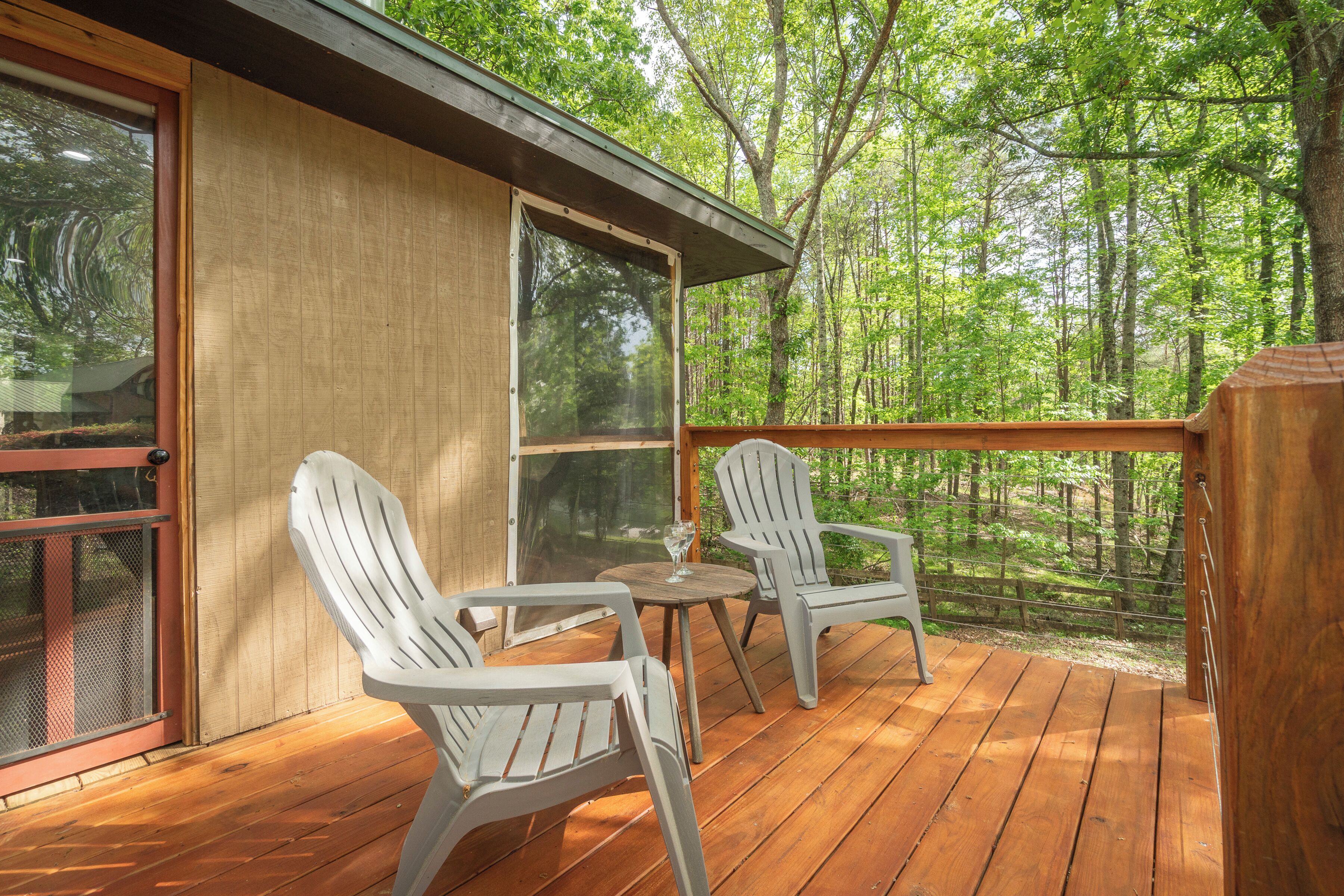 A wooden deck with two white chairs and a table, overlooking a forested area.