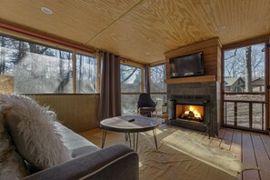 Una sala de estar acogedora con chimenea, un sofá, un televisor y ventanas amplias.