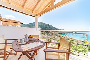 A wooden table with chairs, a white pitcher, and a bowl of fruit on a balcony overlooking a coastal view with mountains in the background.
