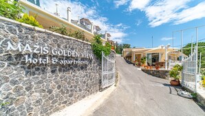 Un mur de pierre sur lequel on peut lire l’enseigne « MAZIS GOLDEN VIEW hotel & apartments », une entrée fermée par un portail et un chemin menant à un bâtiment doté d’un toit en tuiles.