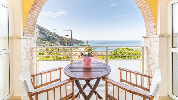 Ein Balkon mit einem Holztisch und -stühlen, einer rosa Vase und Blick auf das Meer und das Grün.