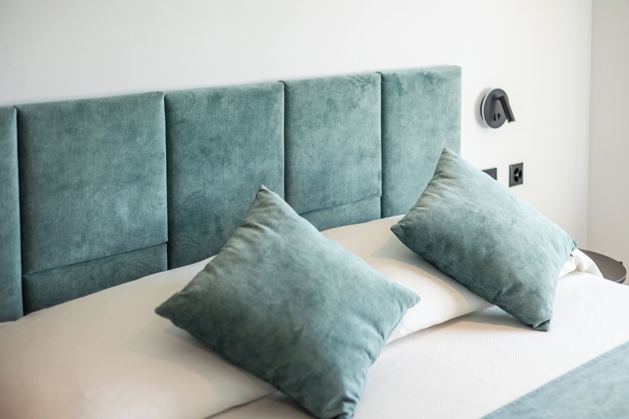 Een bed met een smaragdgroen bekleden hoofdbord, twee passende kussens en een nachtkastje met een lamp.