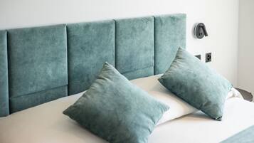 Een bed met een smaragdgroen bekleden hoofdbord, twee passende kussens en een nachtkastje met een lamp.