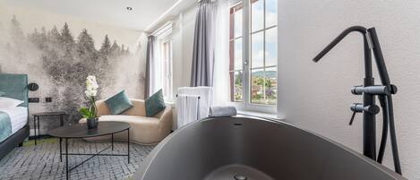 A modern hotel room with a large bathtub, a sofa, a small table with a vase of flowers, and a view of a cityscape through large windows.