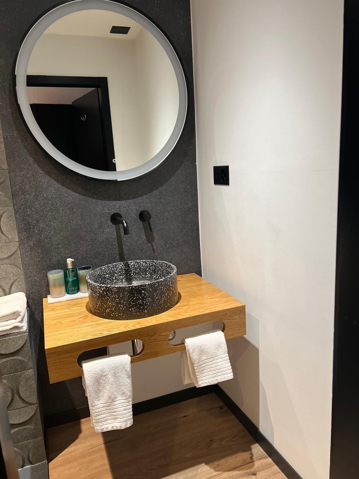 A bathroom with a round mirror, a black speckled sink, and two towels hanging from a wooden cabinet.
