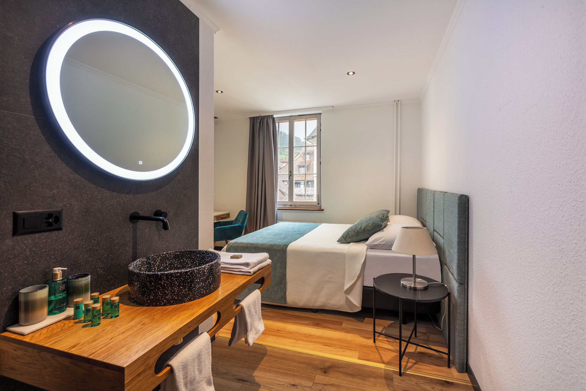 Ein modernes Hotelzimmer mit einem Holzinlay, einem runden Spiegel, einem Bett mit blauer Decke und einem Fenster mit Vorhängen.