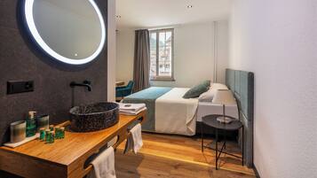 Ein modernes Hotelzimmer mit einem Holzinlay, einem runden Spiegel, einem Bett mit blauer Decke und einem Fenster mit Vorhängen.