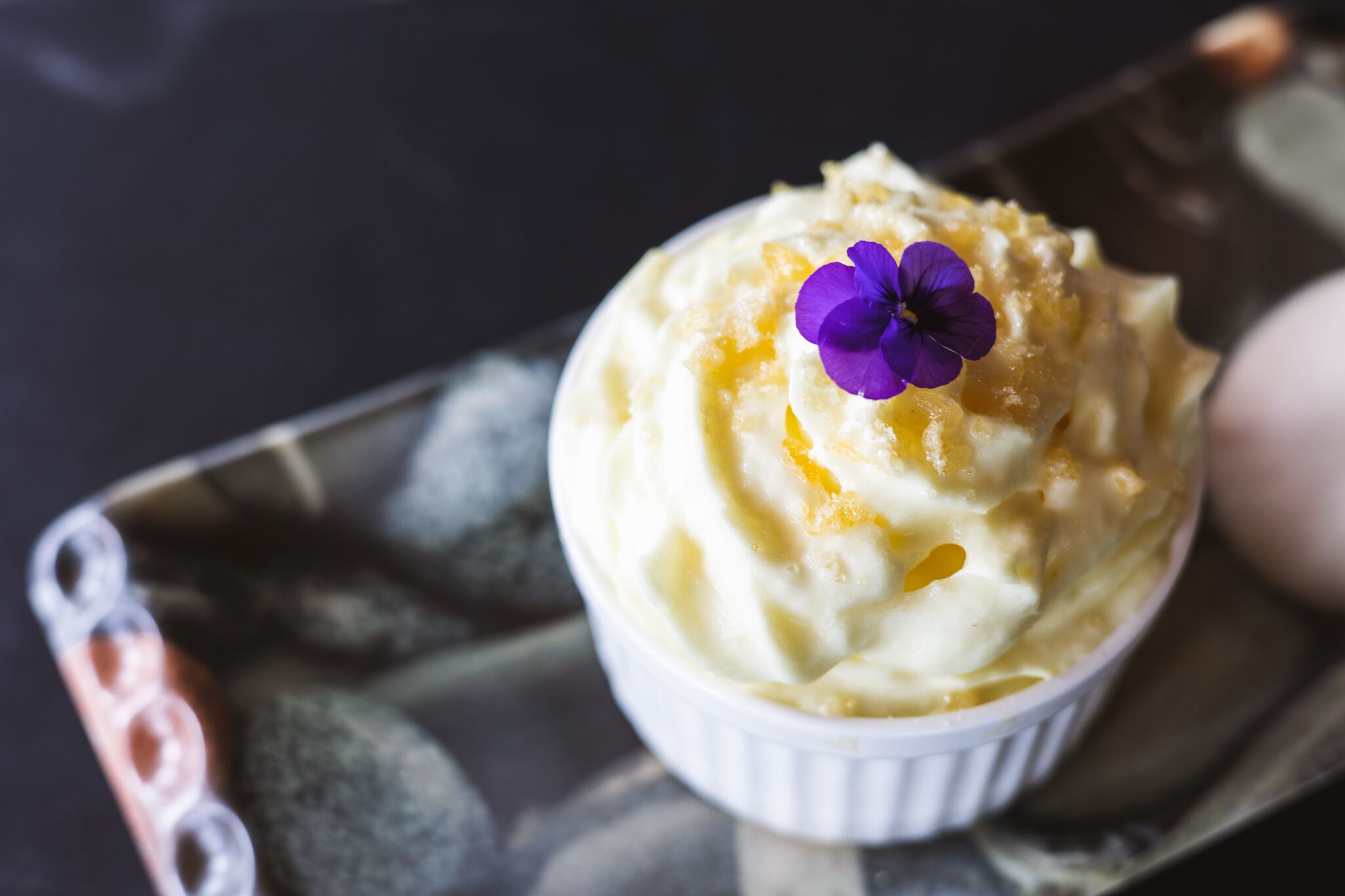 A dessert in a white ramekin on a tray with a purple flower on top.