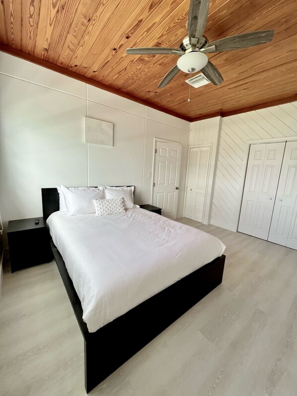 Een slaapkamer met een houten plafond, een bed met wit beddengoed, een nachtkastje en een plafondventilator.
