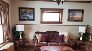 A living room with a brown leather sofa, a wooden coffee table, and a patterned rug. There are two lamps on side tables, a window with curtains, and framed pictures on the wall. - Charming unique historical home on Main street, in the heart of Downtown Bozeman (Bozeman)