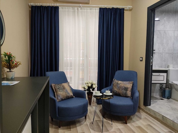 Ein Zimmer mit zwei blauen Sesseln, einem kleinen Tisch mit Tassen, einer Topfpflanze und Blick durch ein Fenster mit blauen Vorhängen.