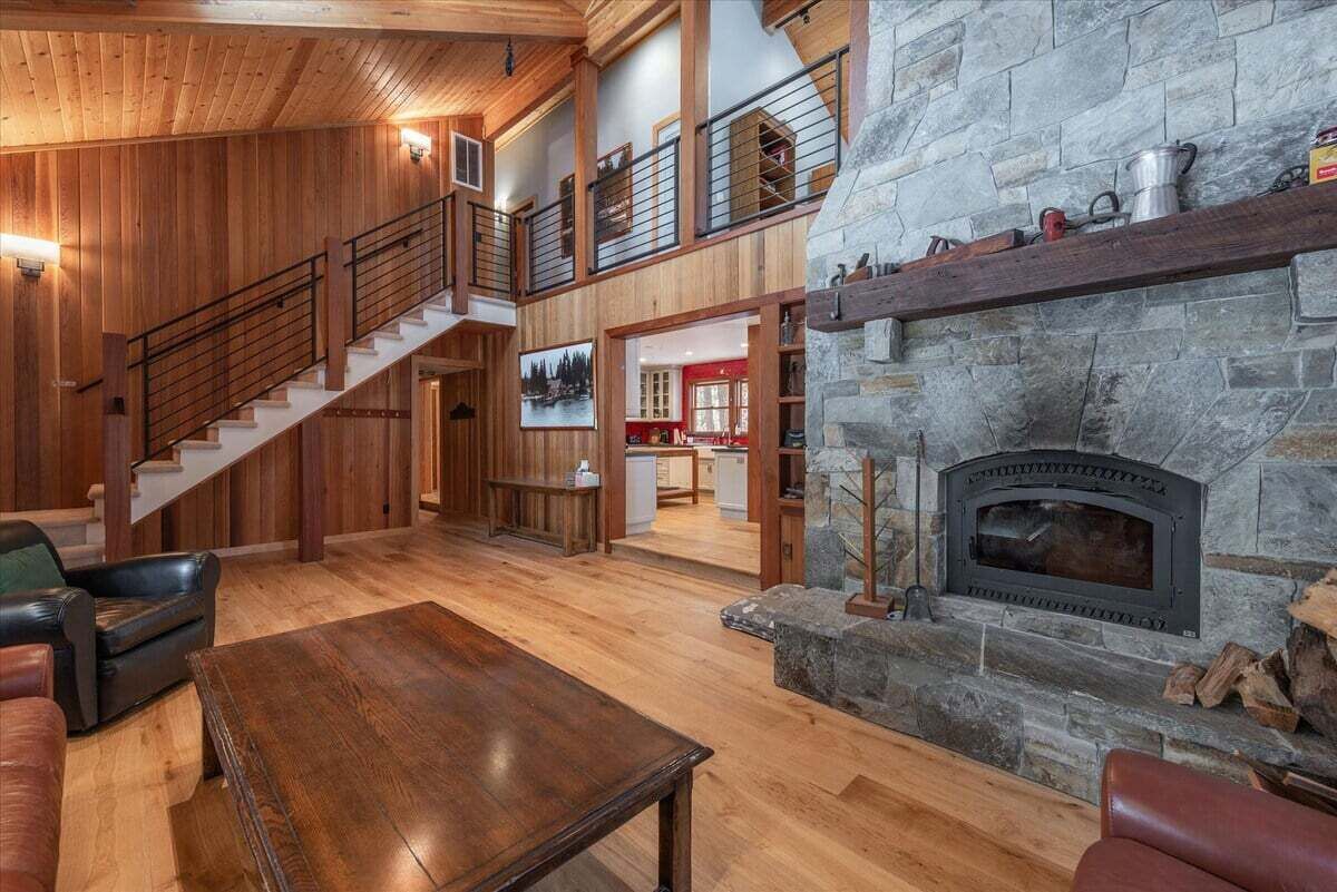 Ein geräumiger Wohnbereich mit Kamin aus Stein, Holzdielen, einer Treppe und einer im Hintergrund sichtbaren Küche.