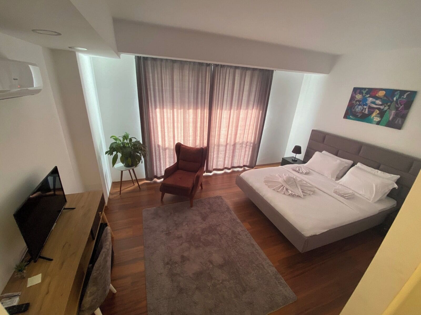 A bedroom with a bed, a chair, a desk, a plant, and a painting on the wall.