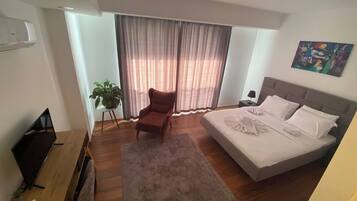 A bedroom with a bed, a chair, a desk, a plant, and a painting on the wall.
