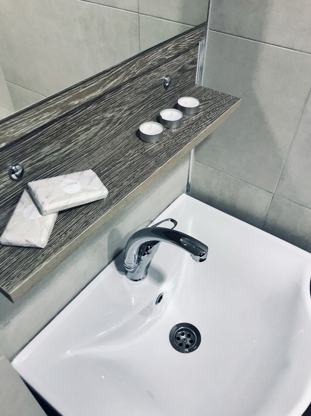 Une salle de bain avec un lavabo blanc, une étagère en bois sur laquelle sont posés du savon et des bougies, et un robinet chromé.