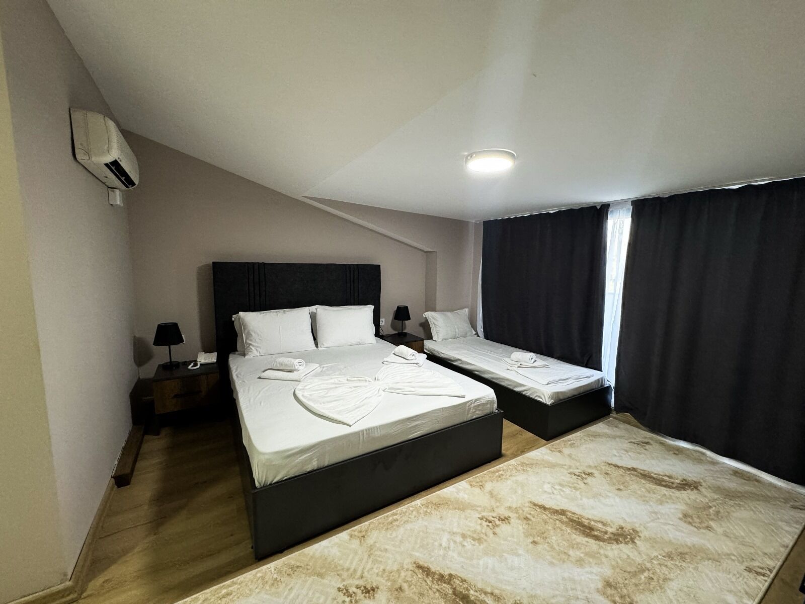 A hotel room with two beds, a ceiling fan, a nightstand with a lamp, and a window with curtains.