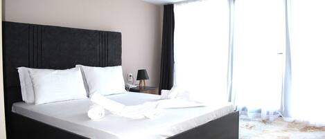 Cama bem arrumada com lençóis e almofadas brancas, cabeceira escura, mesa de cabeceira com abajur e uma janela grande com cortinas.