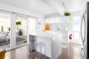 A modern kitchen with a central island, clear glass stools, and a sliding glass door leading to an outdoor area.