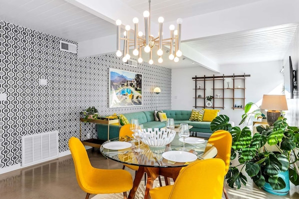 Uma área de refeições com mesa de vidro, cadeiras amarelas, uma tigela decorativa branca e um lustre moderno.