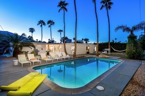 A swimming pool with lounge chairs, palm trees, and a building with a lighted entrance.