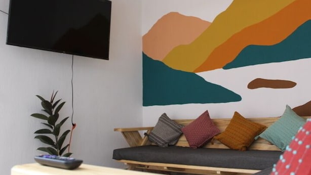 A living room with a flat-screen TV mounted on the wall, a sofa with cushions, a small table with a plant and a remote control, and a wall mural depicting a landscape. - Real 114 Hostal - Hostel (San Cristóbal de las Casas)