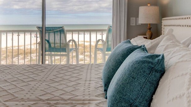 Un dormitorio con un ventanal que da a una playa, una cama con almohadas blancas y azul turquesa, y una silla junto a la ventana.