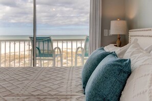Un dormitorio con un ventanal que da a una playa, una cama con almohadas blancas y azul turquesa, y una silla junto a la ventana.