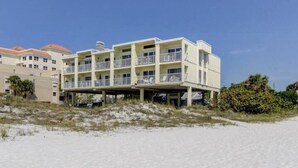 Un hotel de varios pisos con balcones, situado en una playa arenosa con vegetación al fondo.