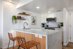 A modern kitchen with a marble island, two wicker bar stools, white cabinets, and a hanging plant. - 429 · Ilikai Collection # 2. Sunset and ocean views! (Honolulu)