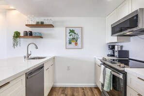 Eine moderne Küche mit weißen Schränken, einer Edelstahl-Spüle, einem eingebauten Backofen und einer Mikrowelle. Darüber befinden sich Regale mit Glasbehältern und einer gerahmten Pflanze.