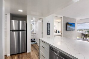 A modern kitchen with a stainless steel refrigerator, a marble countertop, and a view of a bedroom with a beach scene.
