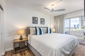 A modern hotel room with a large bed, two bedside lamps, a ceiling fan, and a city view through large windows. - 429 · Ilikai Collection # 2. Sunset and ocean views! (Honolulu)
