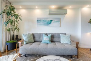 Ein modernes Wohnzimmer mit einem grauen Sofa, zwei gemusterten Kissen, einer Topfpflanze und einer wandmontierten Klimaanlage.