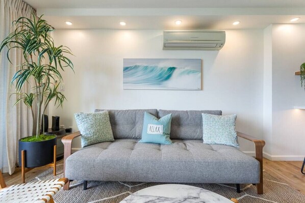 A modern living room with a grey sofa, two patterned cushions, a potted plant, and a wall-mounted air conditioner. - 429 · Ilikai Collection # 2. Sunset and ocean views! (Honolulu)