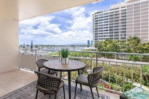 A balcony with a round table and four wicker chairs, overlooking a marina with boats and a cityscape. - 429 · Ilikai Collection # 2. Sunset and ocean views! (Honolulu)
