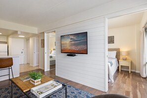 A modern hotel room with a flat-screen TV mounted on the wall, a bed with a bedside table, a small seating area with a coffee table, and a view of the beach through the window. - 1016 · Ilikai Collection # 4. Amazing views, beachfront. (Honolulu)