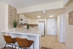 A modern kitchen with white cabinets, a white island with two brown chairs, a microwave, and a refrigerator. - 1016 · Ilikai Collection # 4. Amazing views, beachfront. (Honolulu)