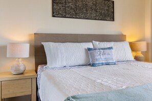 A neatly made bed with a quilted white duvet, two pillows, and a decorative pillow with a blue and white pattern. There are two bedside lamps with round bases on wooden nightstands. - 1016 · Ilikai Collection # 4. Amazing views, beachfront. (Honolulu)