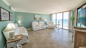 A spacious living room with a large window offering a view of the ocean, a white sofa, two armchairs, a small side table with a lamp, and a decorative wall piece. - Beach Blessings Edgewater 0602T2 Beautiful condo with Beautiful views! (Panama City Beach)
