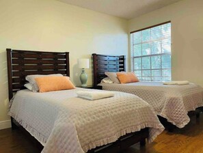 Two single beds with white linens and decorative pillows, a bedside table with a lamp, and a large window.