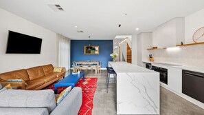 A modern living room with a brown leather sofa, a blue coffee table, and a red rug. There is a dining area with a wooden table and chairs, and a kitchen with white cabinets and a marble countertop.