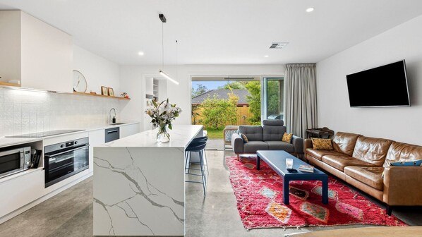 A modern kitchen with a marble island, a dining area with a red rug, a leather sofa, and a TV mounted on the wall.