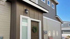 A residential building with a brown front door, a small porch with a chair, and a landscaped area with mulch. - Convenient 3-Bedroom Townhome with Full Garage (Nampa)