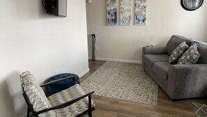 Una sala de estar con un sofá gris, un sillón estampado y un reposapiés azul. Hay cuadros abstractos en la pared y un reloj redondo.