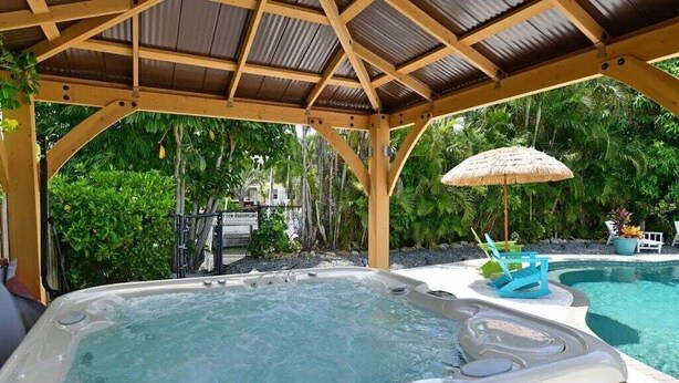 Un jacuzzi bajo una pérgola de madera, con una piscina al fondo.
